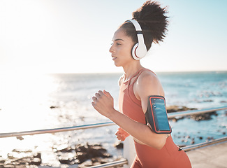 Image showing Black woman, fitness and running with headphones and cellphone on arm at the beach in Cape Town. Sporty African American female runner by the ocean coast having a run for cardio training workout