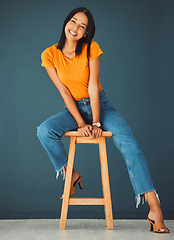 Image showing Black woman, smile and portrait of a young model sitting on a stool in a studio. Casual fashion, happiness and youth of a female feeling relax, calm and happy on a chair isolated by a blue wall