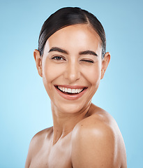 Image showing Portrait, wink and skincare with a model woman in studio on a blue background for natural beauty. Face, skin and winking with an attractive young female posing to promote a luxury cosmetic product