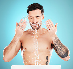 Image showing Water splash, skincare and man cleaning face, morning detox treatment isolated on blue background. Facial hygiene, male model with tattoo and grooming for health, wellness and skin care in studio.