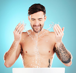Image showing Water splash, sink and skincare of a man cleaning face for wellness, self care and dermatology. Liquid, model ad blue background in a studio with a young man with tattoo doing treatment for beauty
