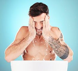 Image showing Water, skincare and man with cleaning face, morning skin treatment isolated on blue background. Facial hygiene, splash and male model with tattoo grooming for health, wellness and beauty in studio.