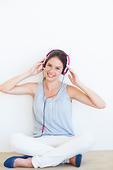 Image showing Portrait, music and woman on a floor with headphones in studio, happy and streaming on a wall background. Face, smile and girl relax with podcast, radio or audio track while sitting against mockup