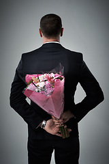 Image showing Valentines day, surprise and flowers of a man in a suit with love present behind back. Flower gift, suit and male model in a isolated gray background studio feeling romantic for a formal date