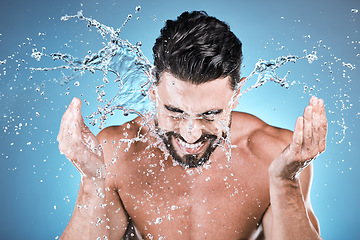 Image showing Water splash, hands and a man model washing his face in studio on a blue background for hygiene or hydration. Bathroom, skincare and cleaning with a handsome young male splashing his skin for beauty