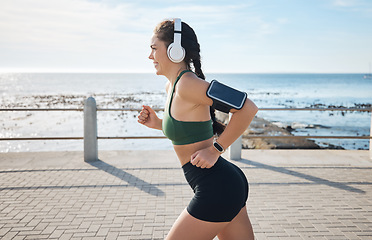 Image showing Music, fitness and woman at beach for running, exercise and morning cardio on blue sky background. Radio, podcast and workout run by girl at ocean for training, happy or relax with audio motivation