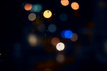 Image showing Bokeh, night and light on a window with water drops, liquid or moisture against a dark abstract background. Blurred lights, colorful and rain drop or splash on glass for city view during rainy season