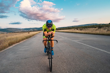 Image showing Triathlete riding his bicycle during sunset, preparing for a marathon. The warm colors of the sky provide a beautiful backdrop for his determined and focused effort.