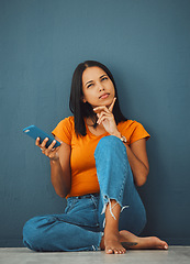 Image showing Phone, thinking and social media with a woman in studio on a blue background, sitting on the floor. Mobile, contact or idea and an attractive young female posing with her finger on her chin