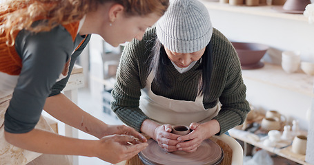 Image showing Pottery teacher, studio and workshop learning, training and teaching to seniorwoman on craft wheel for creativity, clay and sculpture process. Artist helping student in class, design and creative mol