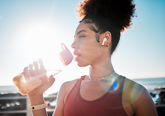 Image showing Drinking water, fitness and black woman on beach exercise, listening to music and cardio training in blue sky lens flare. Liquid bottle for diet, goals and tired sports runner or USA person by ocean