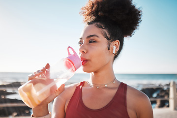 Image showing Fitness black woman drinking water on beach for exercise, listening to music and cardio training in blue sky lens flare. Liquid bottle for diet, goals and tired sports runner or USA person by ocean