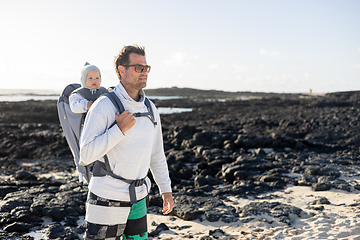 Image showing Young father carrying his infant baby boy son in backpack on black rock volcanic beach on Lanzarote island, Spain. Family travel and winter vacation concept.