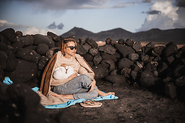 Image showing Mother breast feeding his infant baby boy son on black sandy volcanic beach of Janubio on Lanzarote island, Spain, enjoing dramatic volcanic landscape on windy overcast day. Travel with kids concept