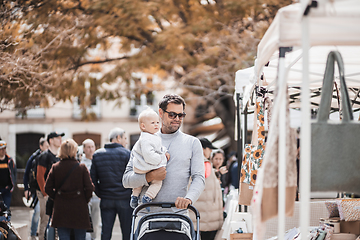 Image showing Father walking carrying his infant baby boy child and pushing stroller in crowd of people wisiting sunday flea market in Malaga, Spain