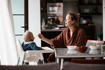 Image showing Cheerful mother wearing bathrope spoon feeding her infant baby boy child sitting in high chair at the dining table in kitchen at home in the morning.