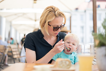 Image showing Young caucasian blonde mother spoon feeding her little infant baby boy child outdoors on restaurant or cafe terrace in summer