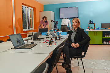 Image showing A woman sitting in a laboratory and solving problems and analyzing the robot's verification. In the background, colleagues are talking at an online meeting