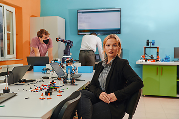 Image showing A woman sitting in a laboratory and solving problems and analyzing the robot's verification. In the background, colleagues are talking at an online meeting