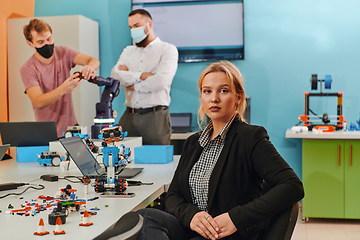 Image showing A woman sitting in a laboratory and solving problems and analyzing the robot's verification. In the background, colleagues are talking at an online meeting