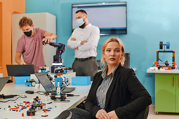 Image showing A woman sitting in a laboratory and solving problems and analyzing the robot's verification. In the background, colleagues are talking at an online meeting