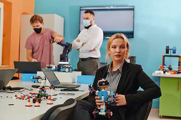 Image showing A woman sitting in a laboratory and solving problems and analyzing the robot's verification. In the background, colleagues are talking at an online meeting