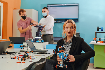 Image showing A woman sitting in a laboratory and solving problems and analyzing the robot's verification. In the background, colleagues are talking at an online meeting