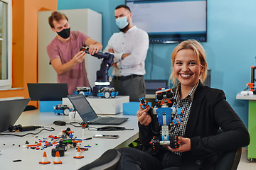 Image showing A woman sitting in a laboratory and solving problems and analyzing the robot's verification. In the background, colleagues are talking at an online meeting