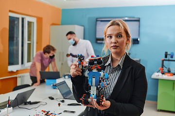 Image showing A woman sitting in a laboratory and solving problems and analyzing the robot's verification. In the background, colleagues are talking at an online meeting