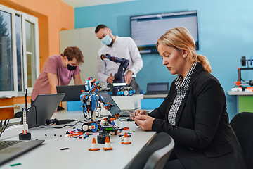 Image showing A woman sitting in a laboratory and solving problems and analyzing the robot's verification. In the background, colleagues are talking at an online meeting