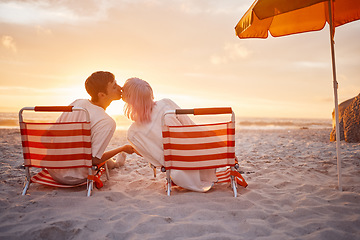 Image showing Couple, kiss and beach chairs by a ocean sunset with love, happiness and care on vacation. Sea, sunshine and kissing of happy young people together sitting by the sand feeling relax outdoor