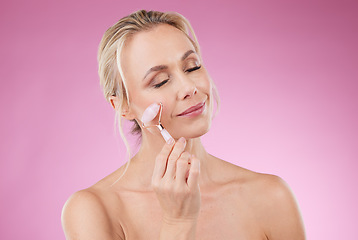 Image showing Beauty, facial roller and woman doing face massage with dermatology and cosmetics in studio. Mature aesthetic person on pink background for skincare, self care and rose quartz results for skin glow