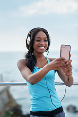 Image showing Black woman, phone and headphones at beach, selfie and smile for video call, music or funny meme to relax. Runner, rest and smartphone with social network post, app and happiness on outdoor adventure