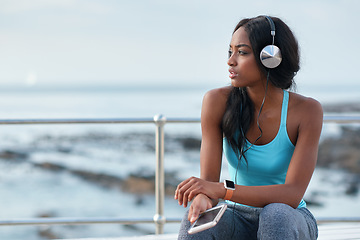 Image showing Black woman, phone and headphones at ocean, sitting and thinking with idea, music and relax to focus. Runner, rest and smartphone with vision, social media app and mindfulness on outdoor adventure