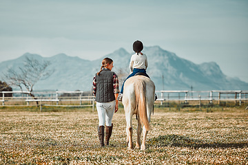 Image showing .Woman leading child on horse, ranch and mountain in background lady and animal walking on field from back. Countryside lifestyle, rural nature and farm animals, mom teaching kid to ride pony in USA.