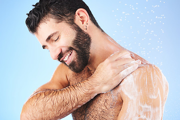 Image showing Shower, cleaning and man with soap, smile and water splash in studio for wellness, hygiene and grooming. Skincare, healthy skin and male with foam, cosmetics and washing body on blue background