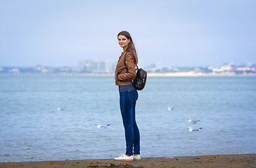 Image showing A girl in autumn clothes with a backpack stands on the seashore, full-length portrait