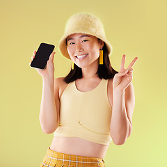 Image showing Portrait, peace sign and Asian woman with phone in studio isolated on a yellow background mockup. Makeup, fashion cellphone and smile of happy gen z female with v hand gesture and mobile smartphone.