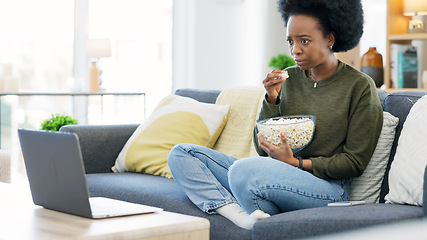 Image showing African woman watching a horror movie on a laptop and eating popcorn while sitting on the couch at home. Terrified black afro female enjoying her online subscription with a variety of scary films