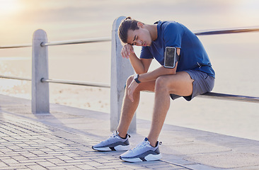 Image showing Tired runner man, sitting and beach for workout, health and exercise with headache, rest and relax. Gen z guy, training burnout and challenge with breathing, mindset and fitness on railing by ocean