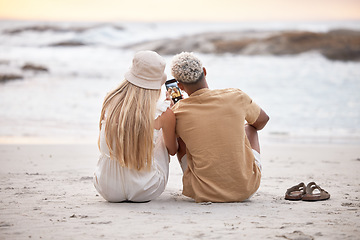 Image showing Rear view of a young couple taking a break and taking a selfie with a smartphone at the beach during sunset. Caucasian female and her mixed race boyfriend relaxing and enjoying the view at the beach