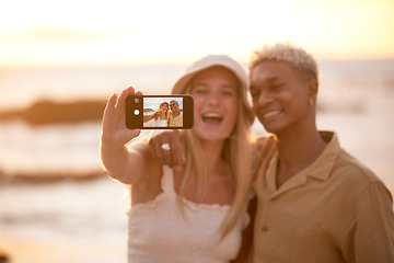 Image showing Closeup portrait of an young affectionate mixed race couple standing on the beach and smiling and taking a selfie with a smartphone during sunset outdoors. Hispanic man and caucasian woman showing l