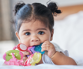 Image showing Portrait, baby girl and biting toys in bedroom, house and home for development, growth and teething progress. Cute kid, face and chewing rattle in mouth for tooth, child and playing in nursery room