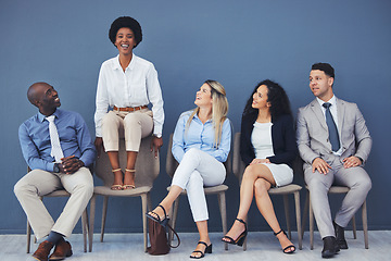 Image showing Black woman, chair and smile with business people for interview with comic laugh, lobby or wall background. Group, we are hiring and waiting room with diversity, happy or opportunity at corporate job