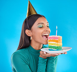 Image showing Celebrate, birthday and woman in studio with a cake, party hat and candle for a celebration. Happy, smile and Indian female model ready to eat sweet rainbow dessert while isolated by blue background.