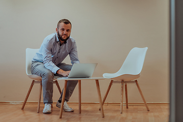 Image showing A confident businessman sitting and using laptop with a determined expression, while a beige background enhances the professional atmosphere, showcasing his productivity and expertise.