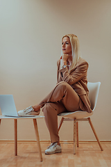 Image showing A professional businesswoman sits on a chair, surrounded by a serene beige background, diligently working on her laptop, showcasing dedication and focus in her pursuit of success