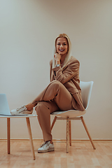 Image showing A professional businesswoman sits on a chair, surrounded by a serene beige background, diligently working on her laptop, showcasing dedication and focus in her pursuit of success