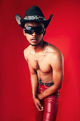 Image showing Fashion, man and cowboy hat, sunglasses and shirtless, clothes and gen z isolated on red background. Confident model, cool and leather pants with eyewear accessory, western aesthetic in a studio