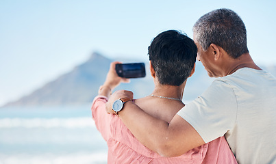 Image showing Beach, photography and back of elderly couple hug in nature for travel, freedom and photo memory. Smartphone, profile picture and rear view of seniors at sea for retirement trip, selfie or vacation